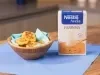 Fotografia em tons claros de uma mesa de madeira com um pano azul. Sobre o pano tem um recipiente de madeira fundo com um paninho azul claro com alguns nuggets saudáveis, ao lado da embalagem de NESTLÉ Farinha de Aveia.
