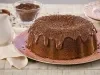 Imagem de um bolo redondo de chocolate com cobertura de brigadeiro e chocolate granulado sobre um prato rosa com bolinhas brancas.