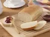 Fotografia em tons de bege em uma bancada de madeira clara, um pano azul colorido em tons pasteis, uma tábua de madeira clara com um pão de forma com fatias e um potinho de geleia ao lado. Ao fundo, uma xícara branca de café e um bule branco.