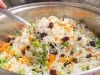 Dentro de uma panela, há arroz branco com uvas-passas, cenouras e ervilhas.