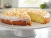 Fotografia de um bolo redondo com crosta dourada e interior amarelo, coberto com glacê branco e flocos de coco tostados, apresentado em um pedestal branco. Uma fatia foi cortada.