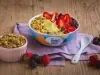 Fotografia em tons de lilás em uma bancada de madeira clara, um bowl colorido com o iogurte de abacate com frutas vermelhas e granola decorando. Ao lado, um potinho com granola e frutas espalhadas na bancada.