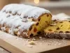Fotografia de um bolo rrecém-assado, colocado em uma tábua de madeira. O bolo, coberto com açúcar de confeiteiro, revela um interior dourado com frutas secas e amêndoas.
