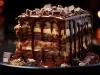 Em cima de uma mesa, prato redondo com fatia de bolo de chocolate, recheado com creme branco e com cobertura de chocolate cremoso.