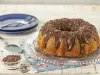 Imagem de um bolo de cenoura com cobertura de brigadeiro e raspas de chocolate sobre um prato redondo transparente.