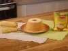 Imagem mostra bolo de milho redondo com furo no meio em um prato vermelho e ao lado de de espigas de milho