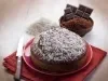 Fotografia de um bolo de chocolate em um prato vermelho, coberto com coco ralado. Ao lado, uma faca serrilhada sobre um guardanapo vermelho, com ingredientes como coco e cacau ao fundo.