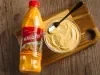 Fotografia em tons de amarelo em uma bancada de madeira, um potinho de vidro com uma mousse de maracujá com uma colher dentro e uma embalagem de um suco de maracujá Maguary ao lado.
