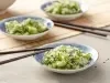 Fotografia em tons de verde em uma bancada de madeira com uma toalha de palha bege. Ao centro, um prato japonês branco com detalhes em azul e a salada de sunomono (pepinos fatiados bem finos) dentro, com um par de hashi apoiado.