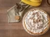 Fotografia vista de cima de uma torta banoffee inteira apoiada em um prato raso branco, sobre uma mesa de madeira.