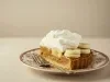 Fotografia em tons claros de uma fatia generosa de banoffee em um prato pequeno e desenhado sobre uma mesa clara. Ao lado da fatia e sobre o prato tem um garfo apoiado.