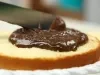 Imagem de um bolo redondo branco cortado com um recheio de brigadeiro sendo espalhada pela espátula.