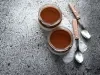 Fotografia vista de cima de dois recipientes de vidro, decorativos, com a sobremesa, finalizada com uma camada grossa de ganache. Ao lado dos recipientes tem duas colheres de sobremesa com o cabo de madeira.