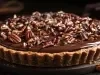 Fotografia em tons mais escuros de uam torta inteira média de uma torta de chocolate com nozes sobre um apoio cinza com nozes espalhadas, ao redor da torta.