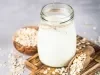Fotografia em tons claros de um pote decorativo com bebida vegetal de coco sobre uma mini tábua de madeira escura retangular. Ao lado do pote tem aveia em flocos espalhadas e dentro de uma colher de madeira longa.