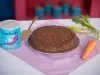 Fotografia de um bolo de cenoura com cobertura de chocolate e ascenda sobre um prato raso grande. Ao redor tem uma cenoura inteira, uma espátula e a alta de ascenda, sobre um pano roxo quadrado.