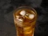 Fotografia em tons escuros de um copo de vidro com uma bebida de guaraná com cubos de gelo, o qual está sobre uma mesa preta.