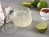 Fotografia em tons de verde em uma bancada cinza com um pano branco e detalhes marrom, uma chaleira de vidro com o chá de limão dentro. Ao lado, uma fatia do limão e um potinho com mel.