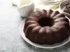 Bolo de chocolate bundt, com cor rica e textura úmida, em um prato branco. Ao fundo, há um bowl com pedaços de chocolate e um jarro de cerâmica.