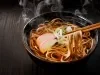 Fotografia mostra um macarrão japonês sendo segurado por hashi, ao fundo um bowl escuro com macarrão dentro.