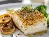 Em cima de uma mesa, prato com posta de bacalhau, cebola caramelizada  e floretes de brócolis.