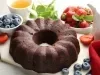 Bolo de chocolate em forma de bundt, com superfície brilhante e densa, servido em uma tábua clara. Ao redor, chá, folhas de hortelã e morangos frescos.