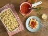 Imagem vista de cima de uma travessa com nhoques, uma panela com molho de tomate, um prato com nhoque com molho de tomate e manjericão e um potinho de queijo ralado.