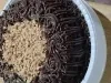Bolo de chocolate com cobertura cremosa em rosetas e crocante granulado no centro, sobre bandeja branca.