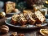 Fotografia de um prato com fatias de bolo de frutas, decorado com açúcar de confeiteiro, cercado por elementos festivos como um raminho de pinheiro, nozes e fatias de laranja seca.