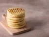 Fotografia vista de cima em tons claros de sete arepas médias, uma sobre a outra, apoiadas em uma mini tábua retangular de madeira, sobre uma mesa clara.