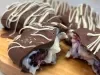 Fotografia de uma tábua de madeira clara sobre uma mesa cinza claro. Sobre a tábua tem alguns bombons coobertos com chocolate meio amargo e decorados com chocolate branco, o recheio é um creme de ninho com frutas vermelhas, um deles está aberto.