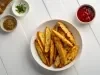 Fotografia em tons claros de um recipiente branco médio, fundo, sobre uma mesa de madeira branca. Dentro do recipiente tem batatas assadas e temperadas, cortadas em meia lua. Ao lado do recipiente tem temperos e ketchup em potinhos brancos.