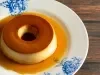 Fotografia de um prato branco, com desenhos azuis, grande raso sobre uma mesa de madeira. Sobre o prato tem um pudim pequeno tradicional feito com amido de milho na receita.
