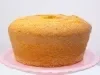 Um bolo de cenoura assado em forma redonda com buraco no meio, colocado sobre um prato rosa. O bolo está dourado por fora, com a superfície levemente rachada, indicando que está bem fofinho.