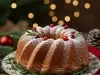 Fotografia de um bolo de Natal em um prato decorativo, com cobertura de açúcar em pó e enfeites de alecrim e cranberries. Ao fundo, uma árvore de Natal com luzes e enfeites vermelhos, criando uma atmosfera festiva.