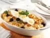 Em cima de uma mesa, refratário com bacalhau, cebola, ovos cortados em rodelas e azeitonas pretas, decorado com folhas de manjericão.