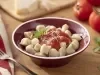 Imagem de um bowl vermelho com nhoque de mandioca com molho de tomate. Ao fundo, tomates, manjericão e um pedaço de queijo parmesão.