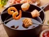 Fotografia em tons claros com uma panela de fondue ao centro. Alguns pedaços de frutos do mar estão espetados no palito para serem mergulhados no fondue