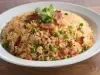 Em cima de uma mesa, prato redondo com porção de arroz soltinho com ervilhas, cenouras picadas, pedaços de bacon, decorado com fatias de bacon e folha de salsa.