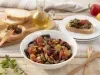 Fotografia em tons de vermelho e amarelo em uma bancada de madeira clara, um prato redondo branco grande ao centro com a caponata de berinjela. Ao lado, uma tábua de madeira com fatias de pães italianos, um potinho de azeite e pimentões amarelo e vermelho