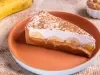 Em cima de uma mesa, prato redondo com fatia de banoffee com massa de biscoitos crocante, doce de leite, bananas em rodelas, decorado com chantilly cremoso e polvilhado com canela em pó.