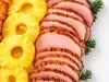 Fotografia em tons claros de faias de tender sobrepostas, deitadas e em fileira, com alguns galhos verdes de natal. Ao lado das fatias tem várias rodelas de abacaxi, também sobrepostas.