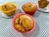 Fotografia de três forminhas de cupcake com bolo de cenoura e uva passa, sendo duas de tom rosa e uma amarela. Duas delas estão sobre um miniprato cinza raso, os quais estão sobre uma toalha de mesa de tom azul claro com bolinhas pretas.