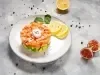 Fotografia em tons claros de uma mesa de cimento queimado claro. Sobre a mesa tem um prato branco raso com tartar de salmão sobre abacate em cubos ao lado de duas rodelas de limão siciliano.