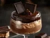 Fotografia em tons escuros de um recipiente de vidro, como uma taça, com camadas de creme e bolacha, cobertas por chocolate.