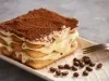 Fotografia em tons claros de uma mini tiramisú clássica sobre um apoio retangular claro, ao lado tem alguns grãos de café inteiros e um garfo apoiado. O recipiente está sobre uma mesa cinza.