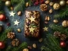Fotografia de um bolo inglês decorado para as festas, com frutas secas, nozes e estrelas douradas, cercado por ramos de pinheiro e enfeites natalinos.