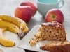 Fotografia de uma fatia de bolo de frutas em um prato branco, acompanhada de bananas e maçãs. O cenário é casual e acolhedor, com uma xícara azul ao fundo, sugerindo um lanche caseiro.