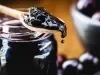 Fotografia de uma colher de madeira sobre um recipiente de vidro com geleia de jabuticaba. A colher também carrega um pouco da geleia, que cai aos poucos na ponta.