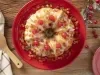 Imagem vista de cima do bolo natalino sobre um prato vermelho decorando com glacê branco, frutas cristalizadas e cerejas.